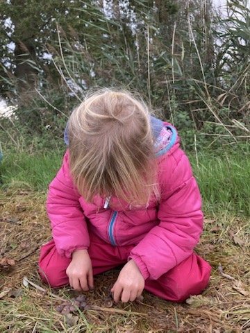 Een kind in een roze jas zit geknield in het gras en onderzoekt de grond