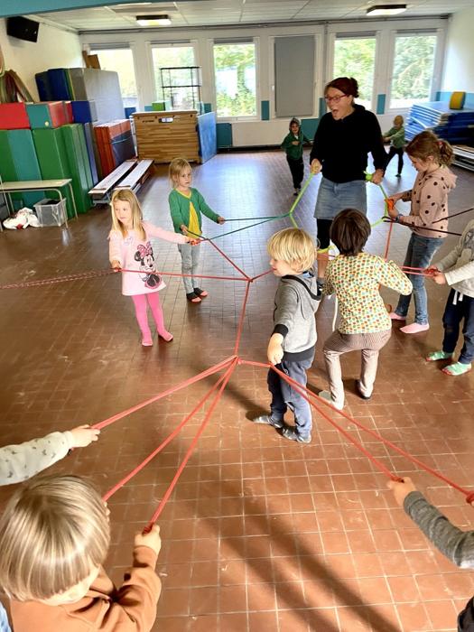 Mieke en kinderen houden samen een groot web van touwen vast in een turnzaal
